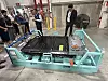 A large battery pack assembly sits on a turquoise metal frame in an industrial workshop with several people standing nearby.